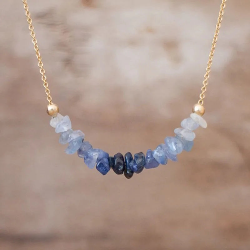 A gold chain necklace featuring a curved row of raw, irregular blue gemstone chips, with a central cluster of dark navy stones and gold bead accents on either side, set against a soft, blurred brown background.