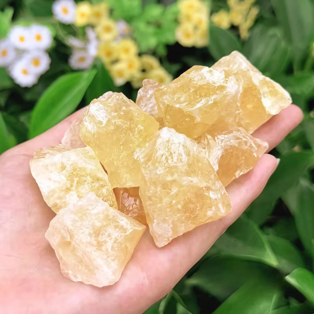A hand holding several rough, translucent yellow crystals against a blurred background of green leaves and small white and yellow flowers.