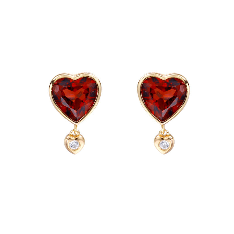 A pair of gold heart stud earrings featuring a large faceted red heart-shaped gemstone on the top and a small dangling heart with a clear round gemstone below, set against a plain white background.