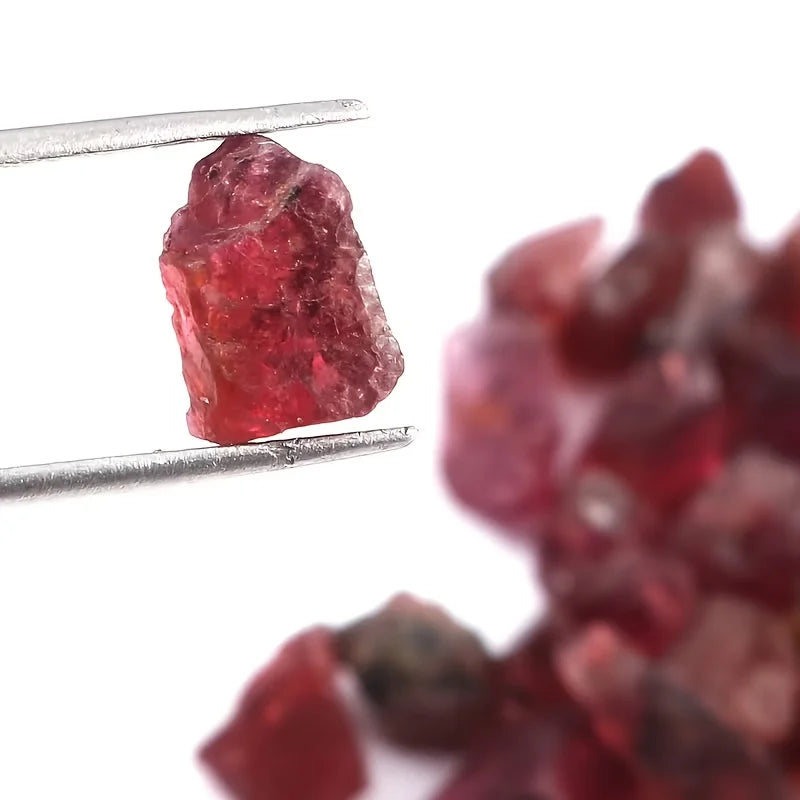A rough, chunky red gemstone held between metal tweezers against a white background, with a blurred pile of similar red gemstones in the foreground.