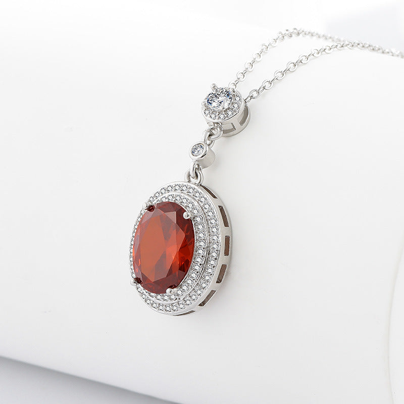 A silver pendant necklace featuring an oval faceted red gemstone surrounded by a halo of small clear stones, suspended from a delicate silver chain on a white background.