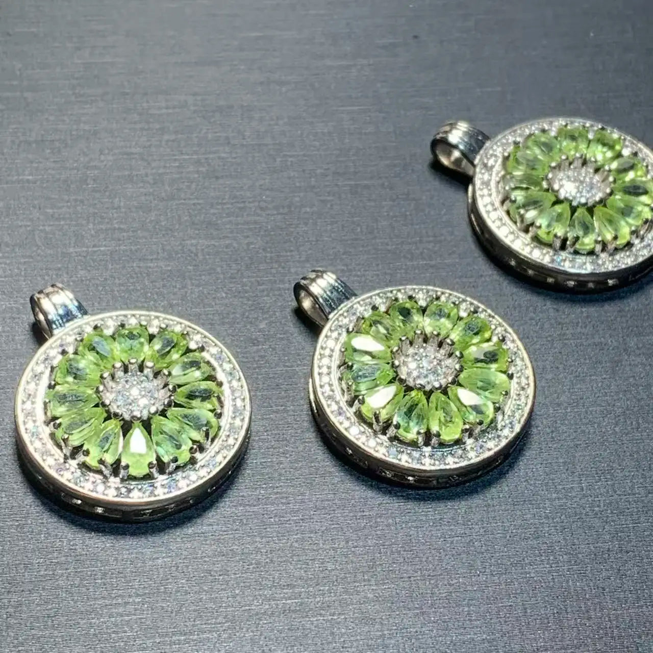 Three round silver pendant charms featuring a green gemstone floral pattern with a clear central stone, displayed on a dark gray textured surface.