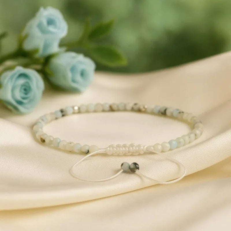 A delicate beaded bracelet with pale blue-green and white beads, featuring a white elastic cord with a small dark bead accent, displayed on draped cream satin fabric with blurred light blue roses in the background.