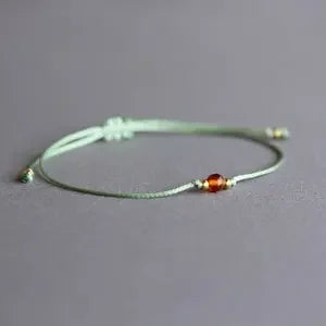 A delicate light green cord bracelet with a small amber-orange bead and tiny gold-toned accents, displayed on a neutral gray background.