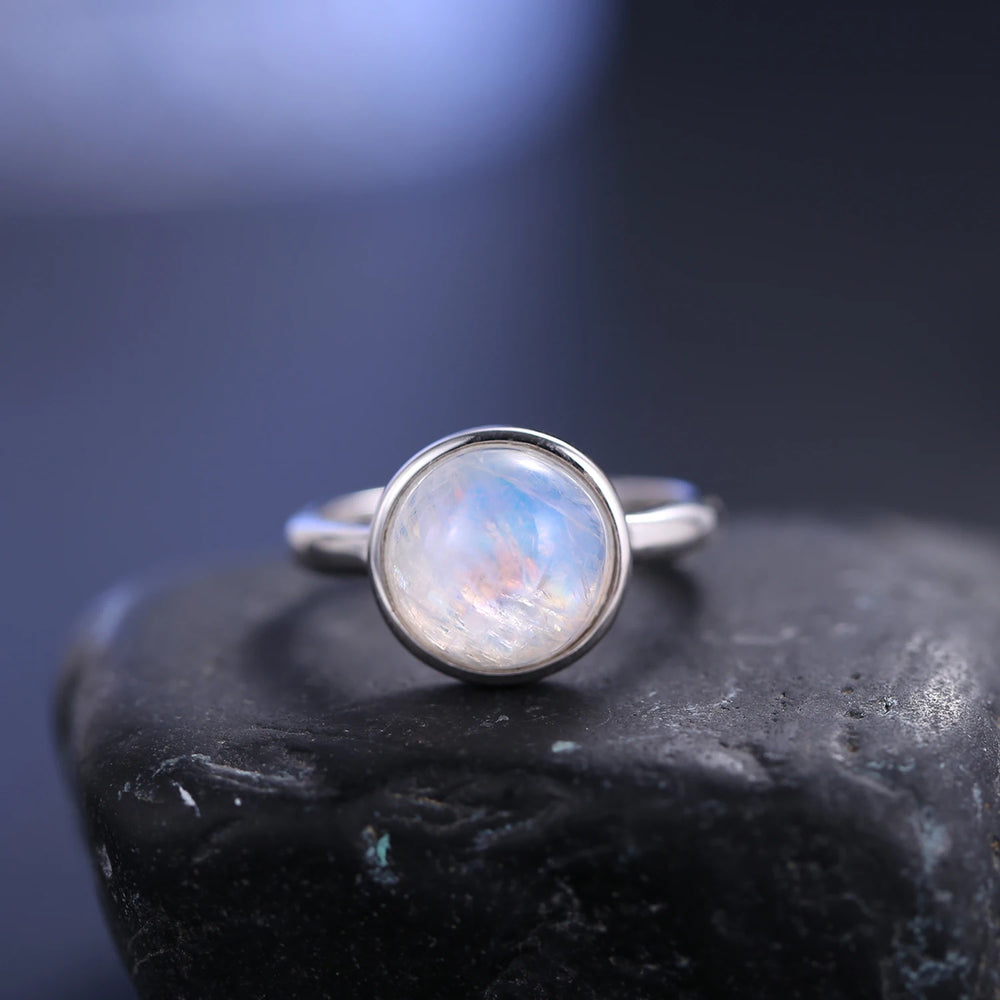 A silver ring with a round iridescent moonstone cabochon, resting on a dark textured stone against a dark blue-gray background.